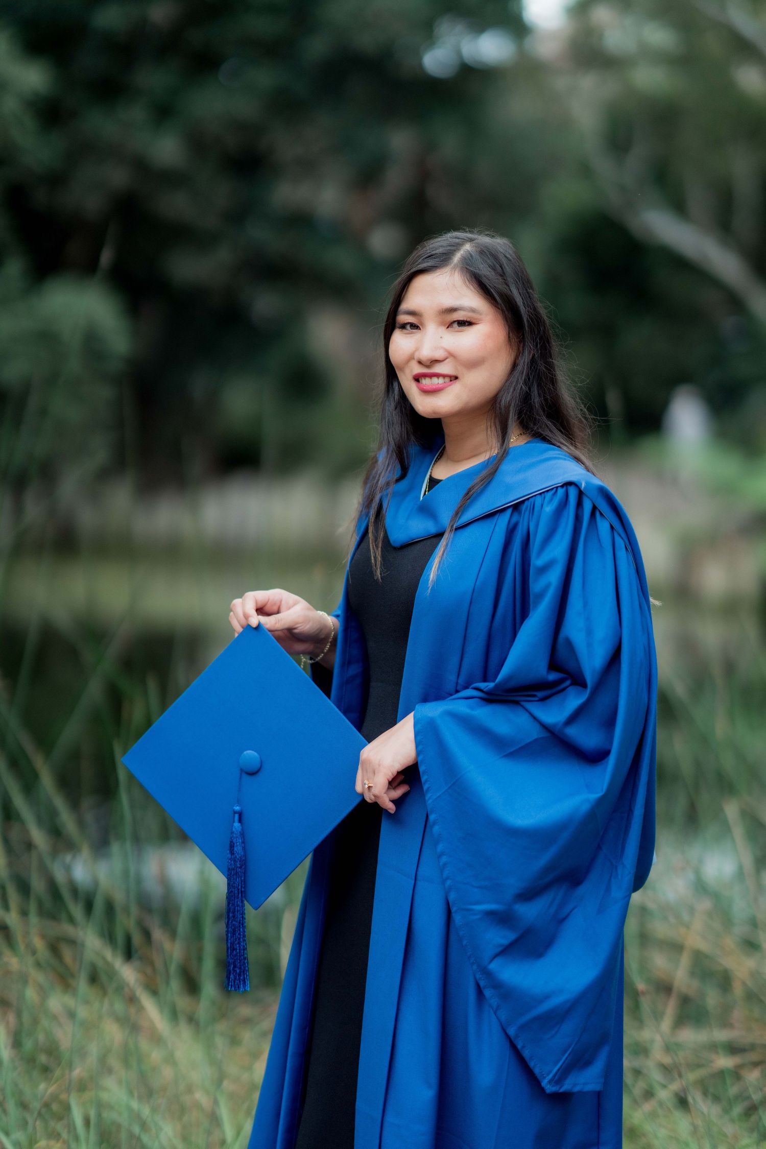 Graduate in a blue gown smiles while holding her mortarboard cap with tassel in a green outdoor setting.