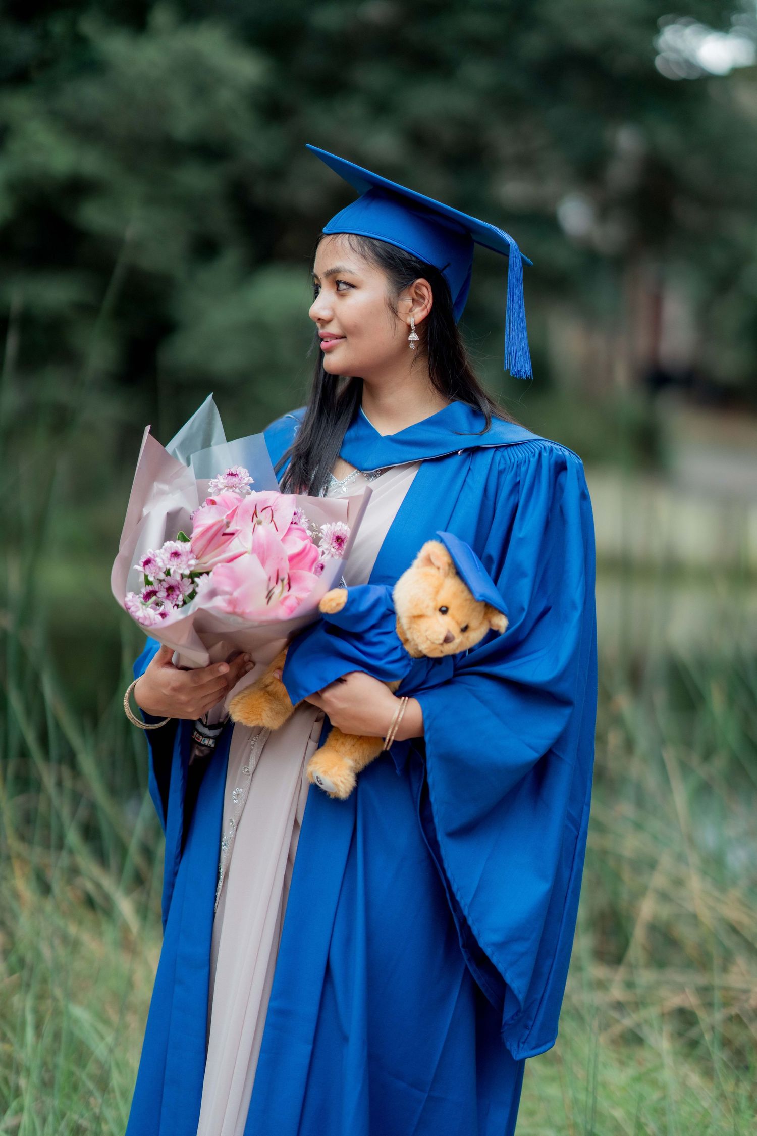 Graduate in a blue cap and gown holds a bouquet of pink flowers and a teddy bear in a graduation outfit while standing outdoors by greenery.
