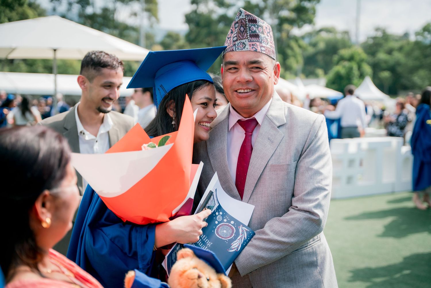 Smiling graduate in a blue cap and gown holds flowers and a teddy bear while posing with a man in a gray suit and traditional hat.