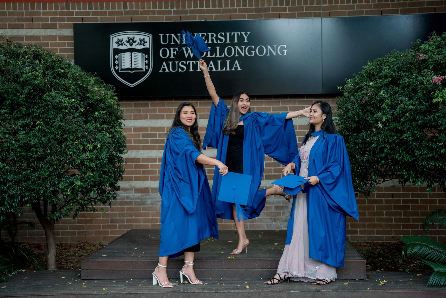 Three graduates in blue gowns toss caps and pose playfully in front of the University of Wollongong Australia sign.