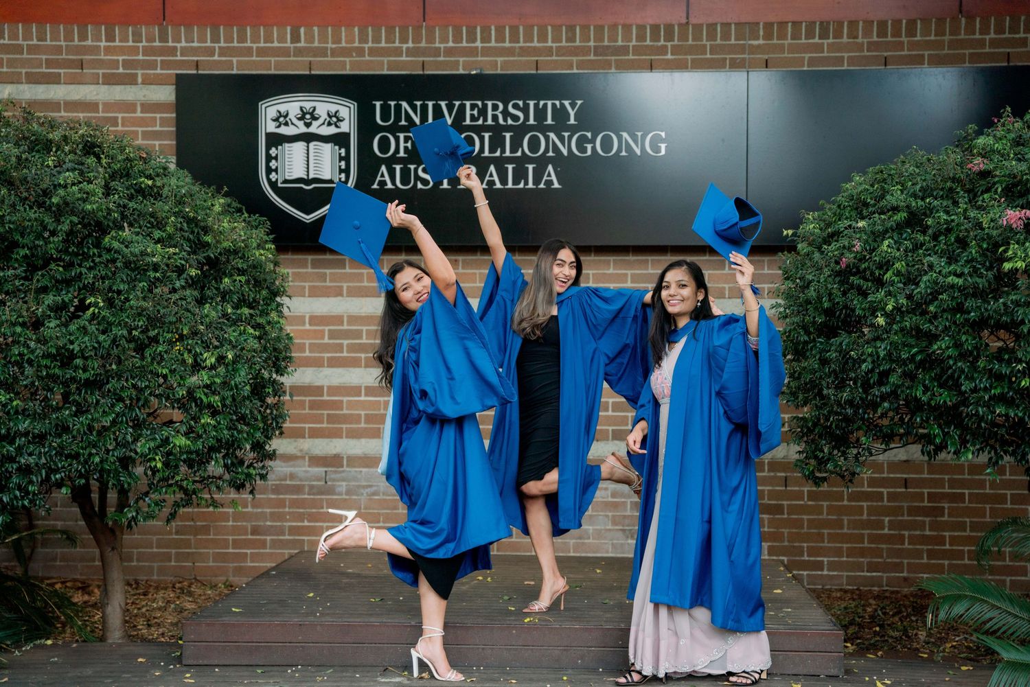 Three female graduates in blue gowns celebrate by raising caps and kicking up heels at the University of Wollongong sign.