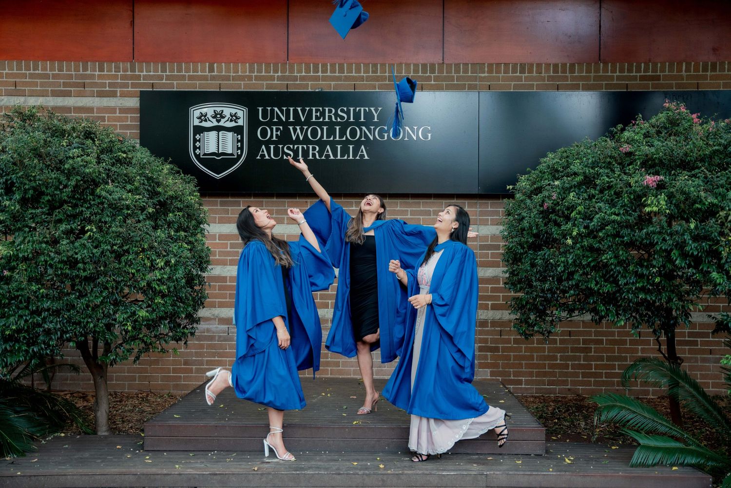 Three graduates in blue gowns celebrate and toss caps in the air outside the University of Wollongong Australia sign.