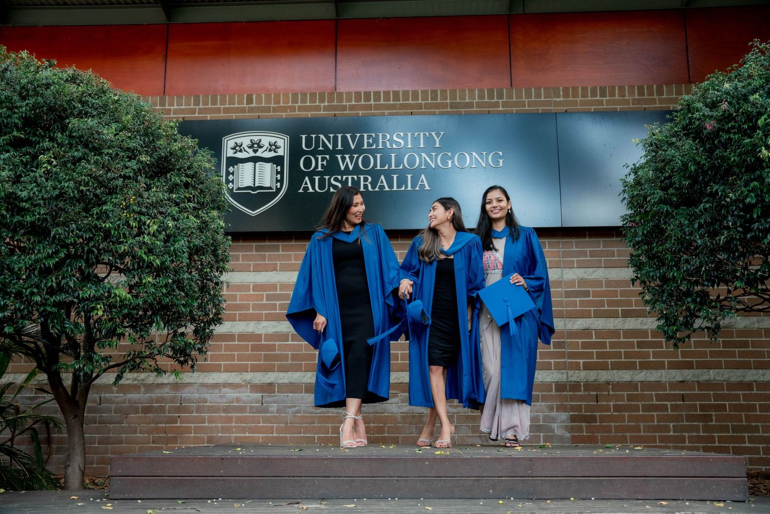 Three University of Wollongong Australia graduates in blue gowns and caps walk arm in arm down brick steps beneath the university sign.