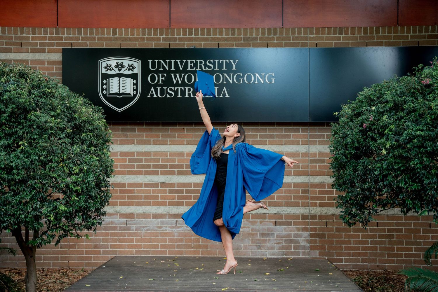 Graduate in blue gown strikes a joyful dance pose with raised arm in front of the University of Wollongong Australia sign.