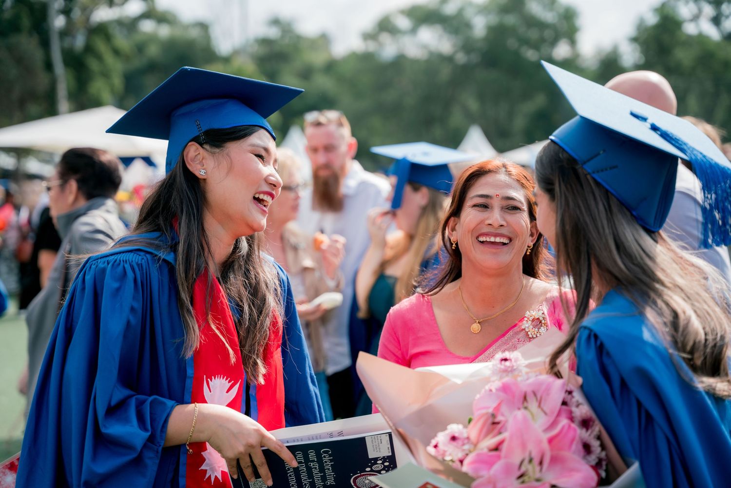 Smiling graduates in blue caps and gowns celebrate at an outdoor graduation ceremony while a woman holds a pink flower bouquet.