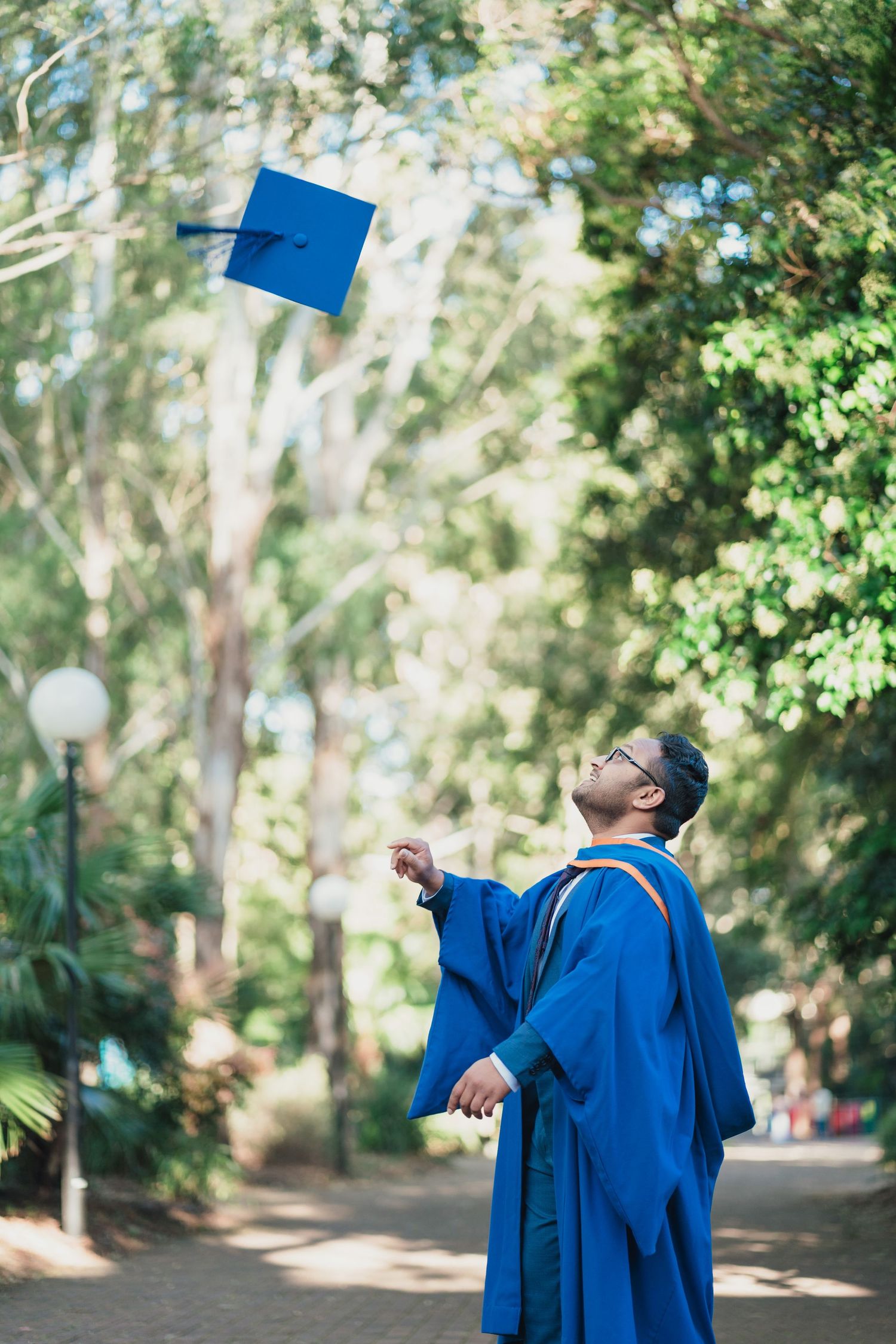 Graduate in a blue gown and glasses tosses a blue mortarboard into the air on a tree-lined path, celebrating graduation day.