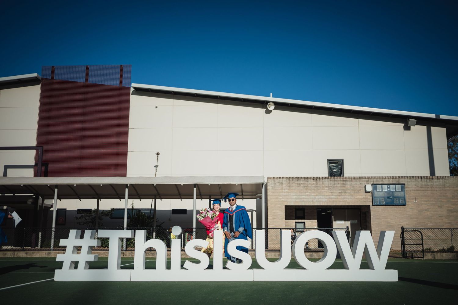 Two people pose behind large white #ThisIsUOW letters outside a campus building under a bright blue sky.