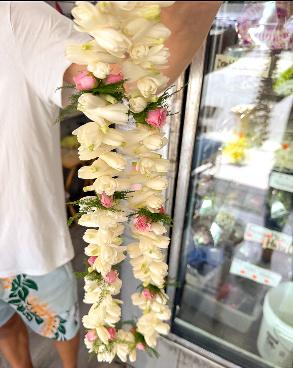 Person holding a long white tuberose flower lei with pink rosebuds and green leaves outside a flower shop display cooler.