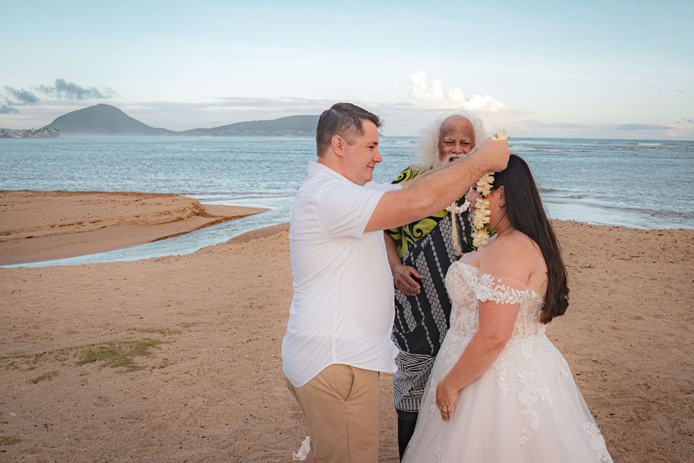Groom places a flower lei on a bride in a white lace gown during a beachfront wedding ceremony led by an officiant at sunset.