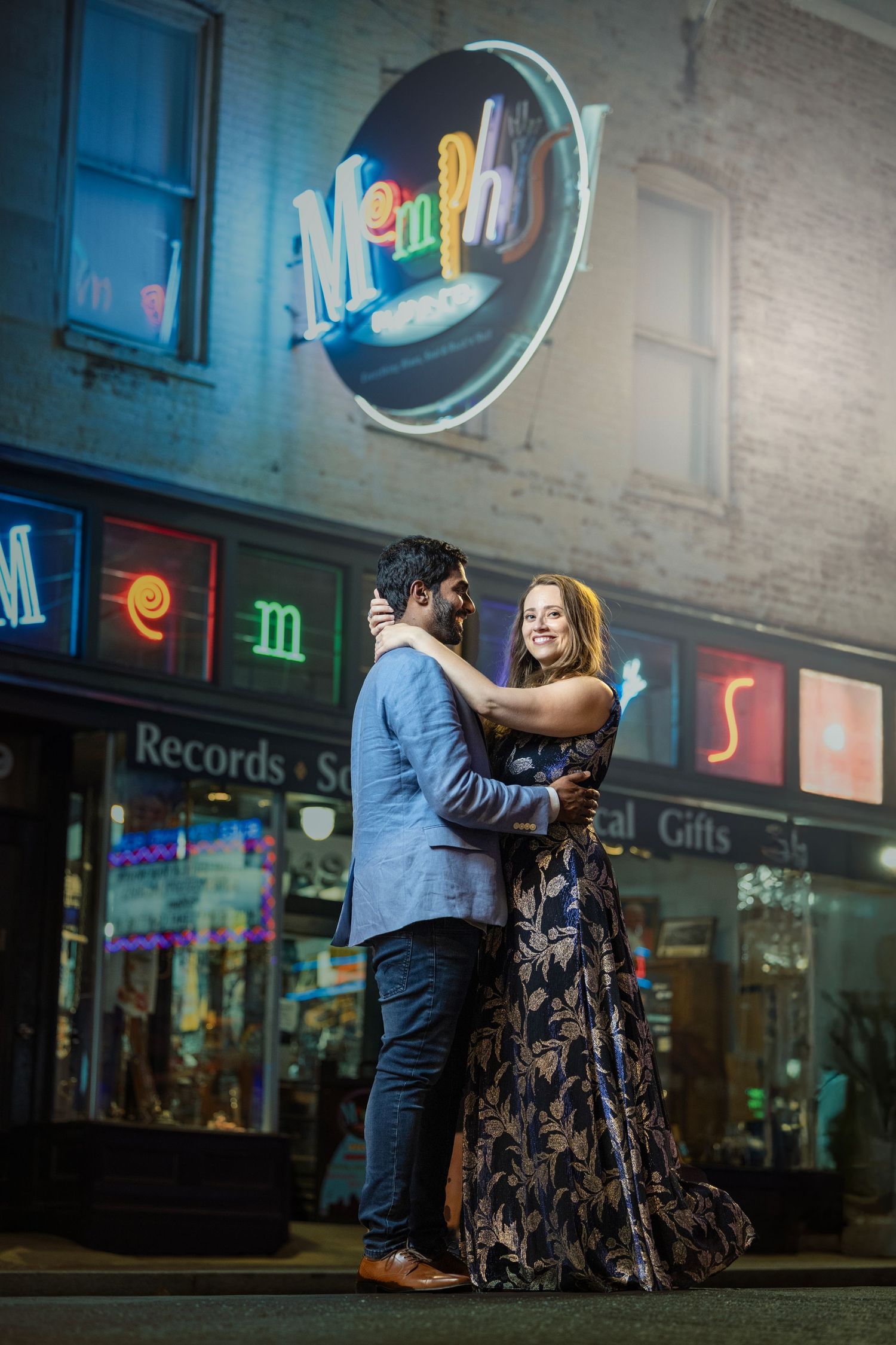 Couple embracing in an evening engagement photo on a city street beneath a glowing Memphis neon sign and colorful record shop storefront.