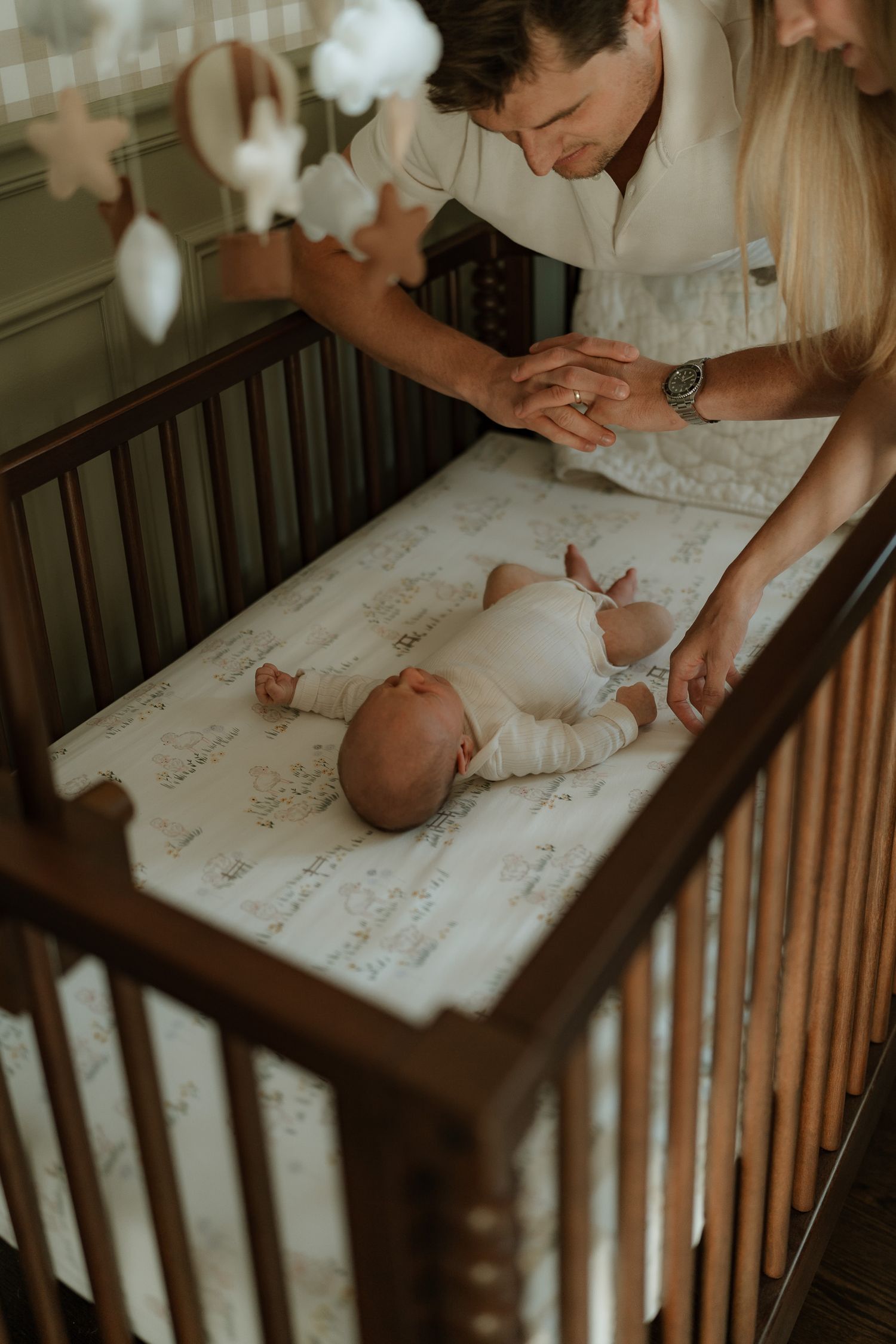 Two parents hold hands over a sleeping newborn baby lying in a dark wood crib with a printed sheet and a star mobile above.