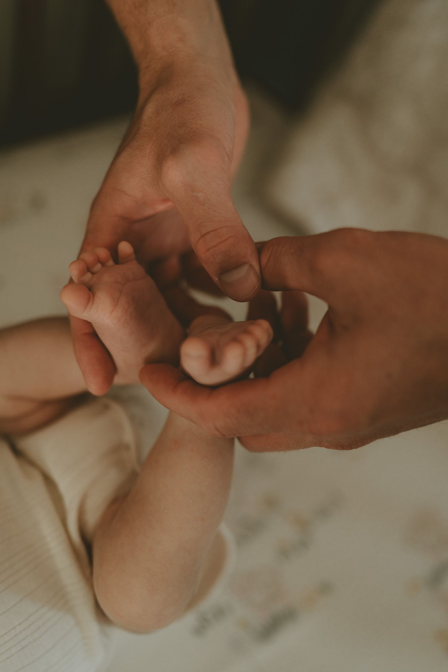 Two adult hands gently cradle the tiny bare feet of a newborn baby lying on a soft, light-colored blanket.