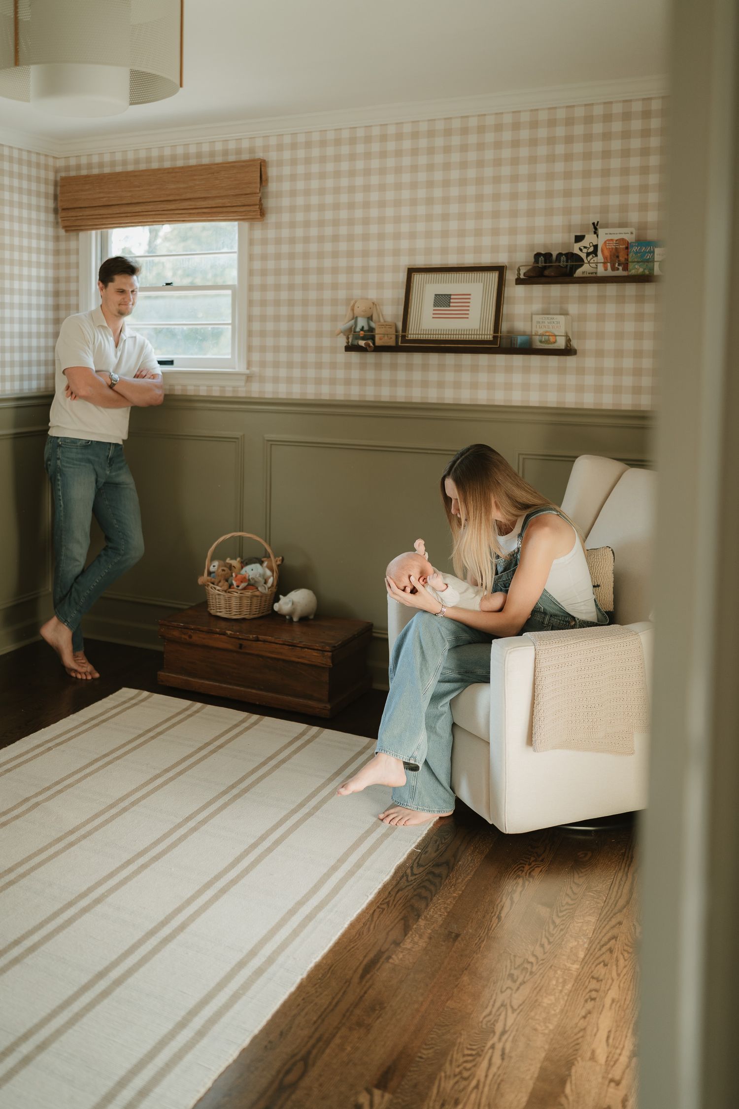 A woman sits in a white armchair holding her newborn baby while a man leans against the wall in a cozy nursery with gingham wallpaper.