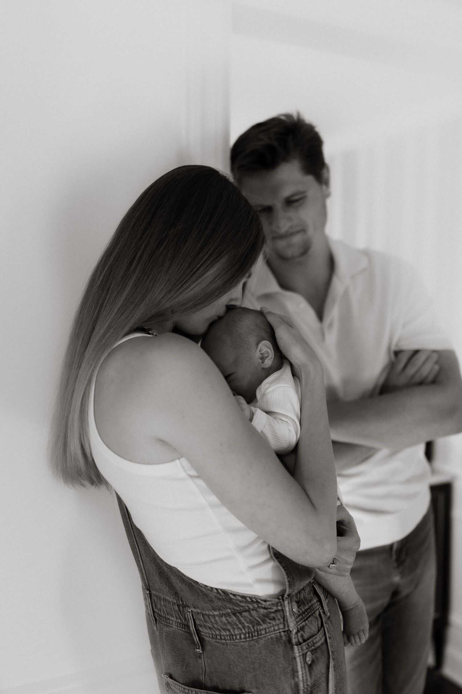 Black and white newborn family portrait of a mother cradling her sleeping baby while the father stands behind with arms crossed.