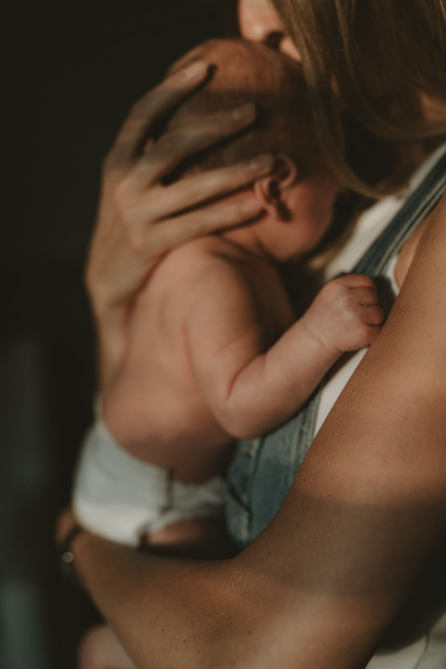 Mother in a denim apron holding a bare-chested newborn baby close to her shoulder in warm, moody natural light.