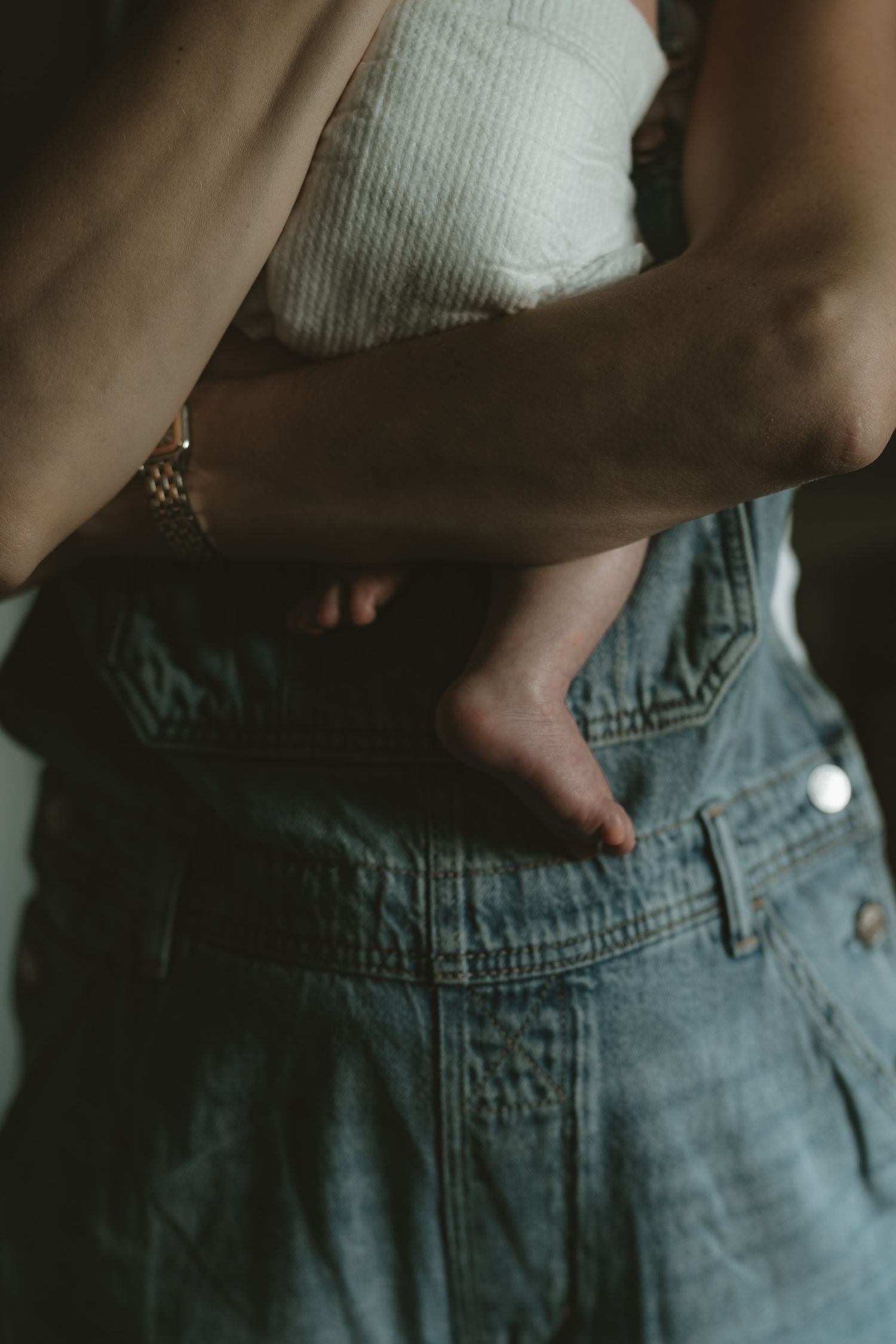 Woman in denim overalls and a gold watch cradles a baby in a diaper against her chest in soft, moody light.