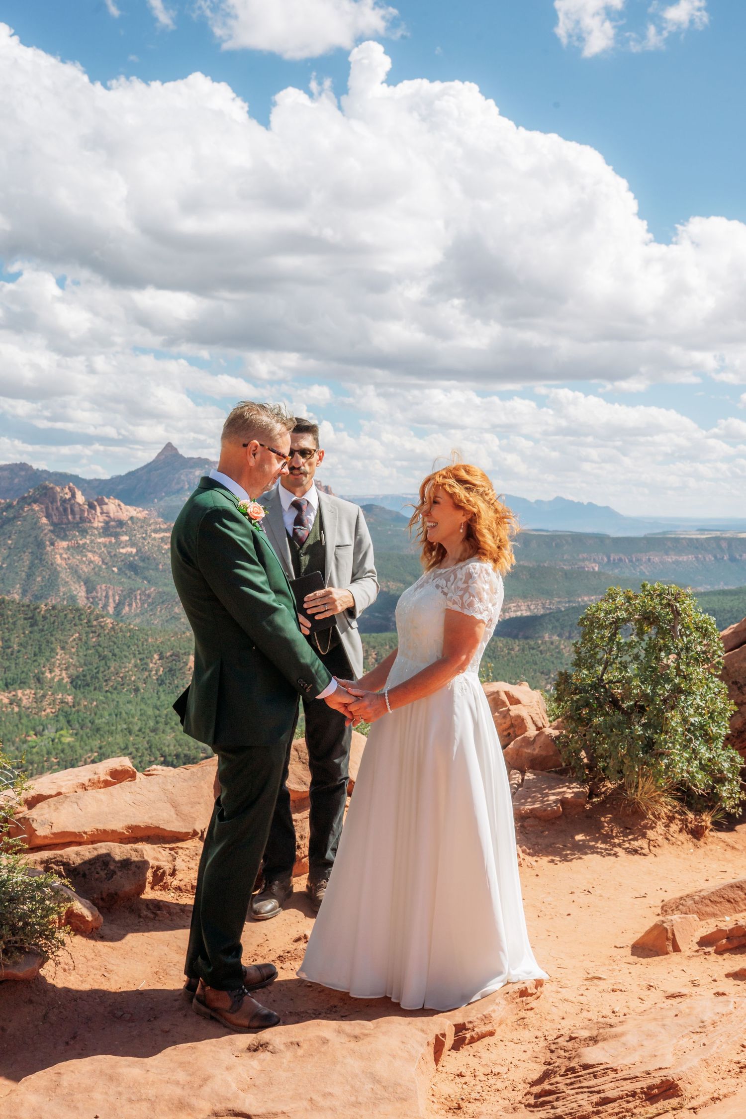 Couple holding hands during an outdoor red rock elopement ceremony with an officiant overlooking a scenic canyon under a cloudy blue sky.