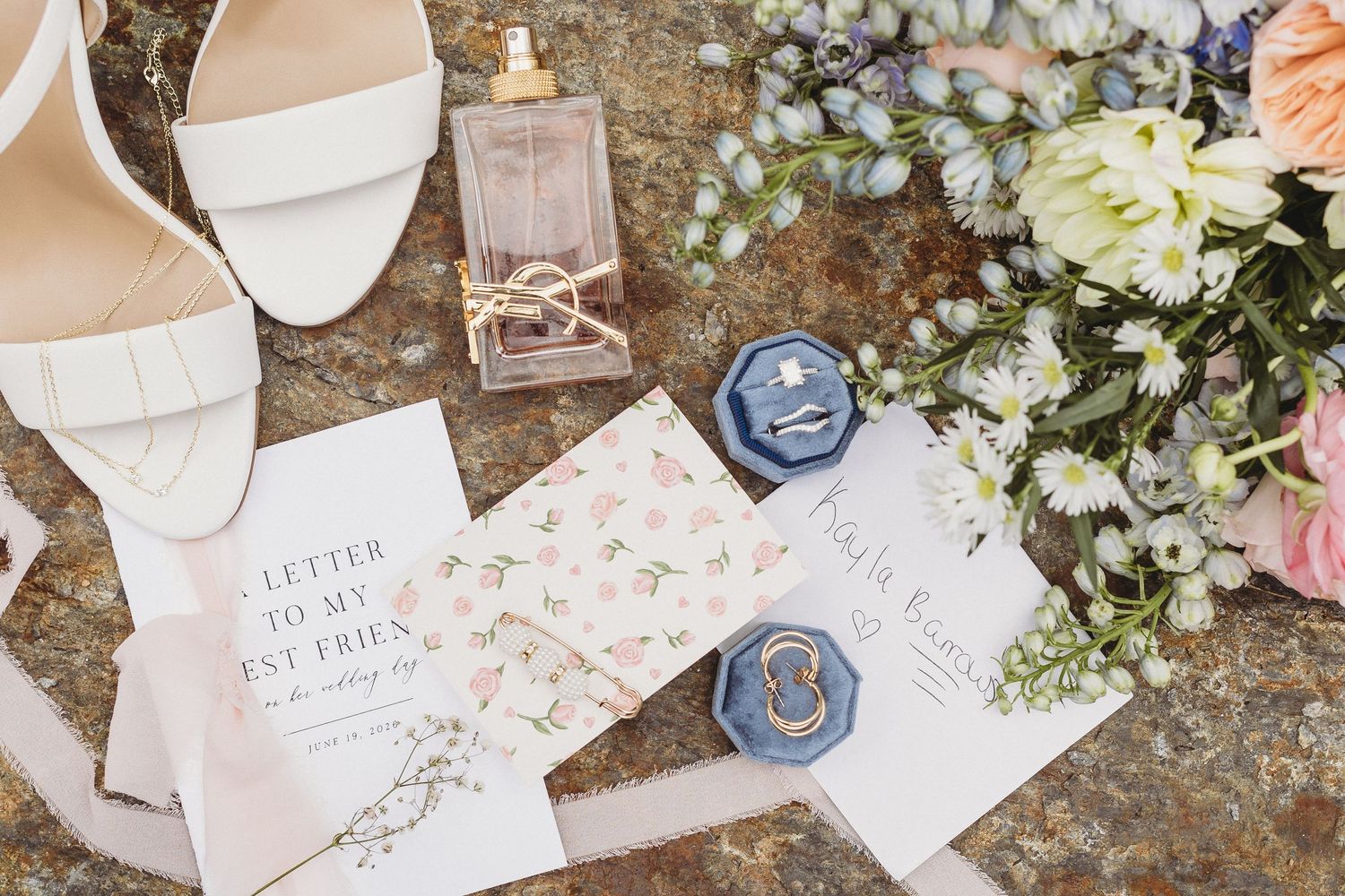 Overhead wedding detail photograph of bridal shoes, perfume, rings in blue velvet boxes, vow books, and pastel flowers.
