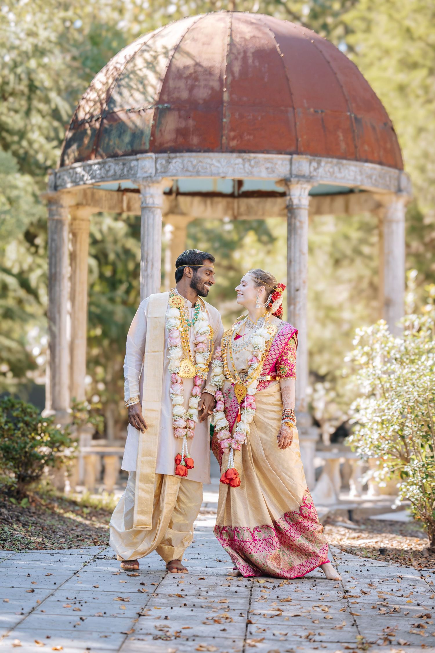 Couple in traditional Indian wedding attire with floral garlands smiling at each other in front of a historic stone gazebo.