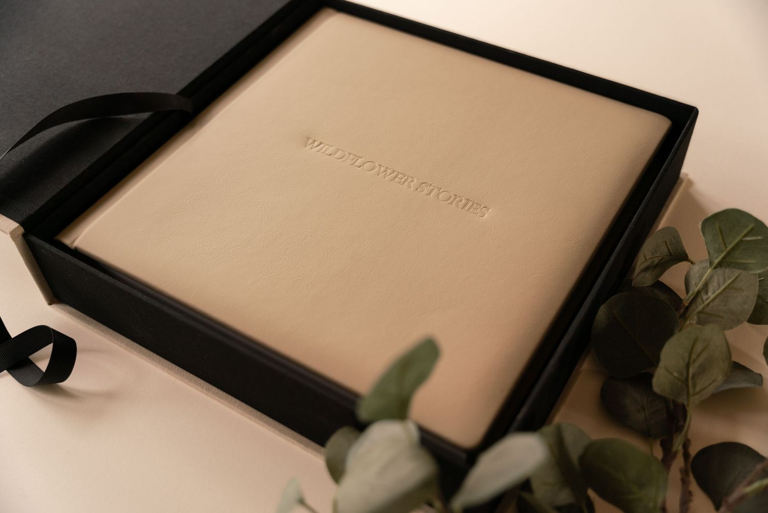 Cream leather photo album with embossed lettering in a black presentation box with ribbon, surrounded by green eucalyptus leaves.