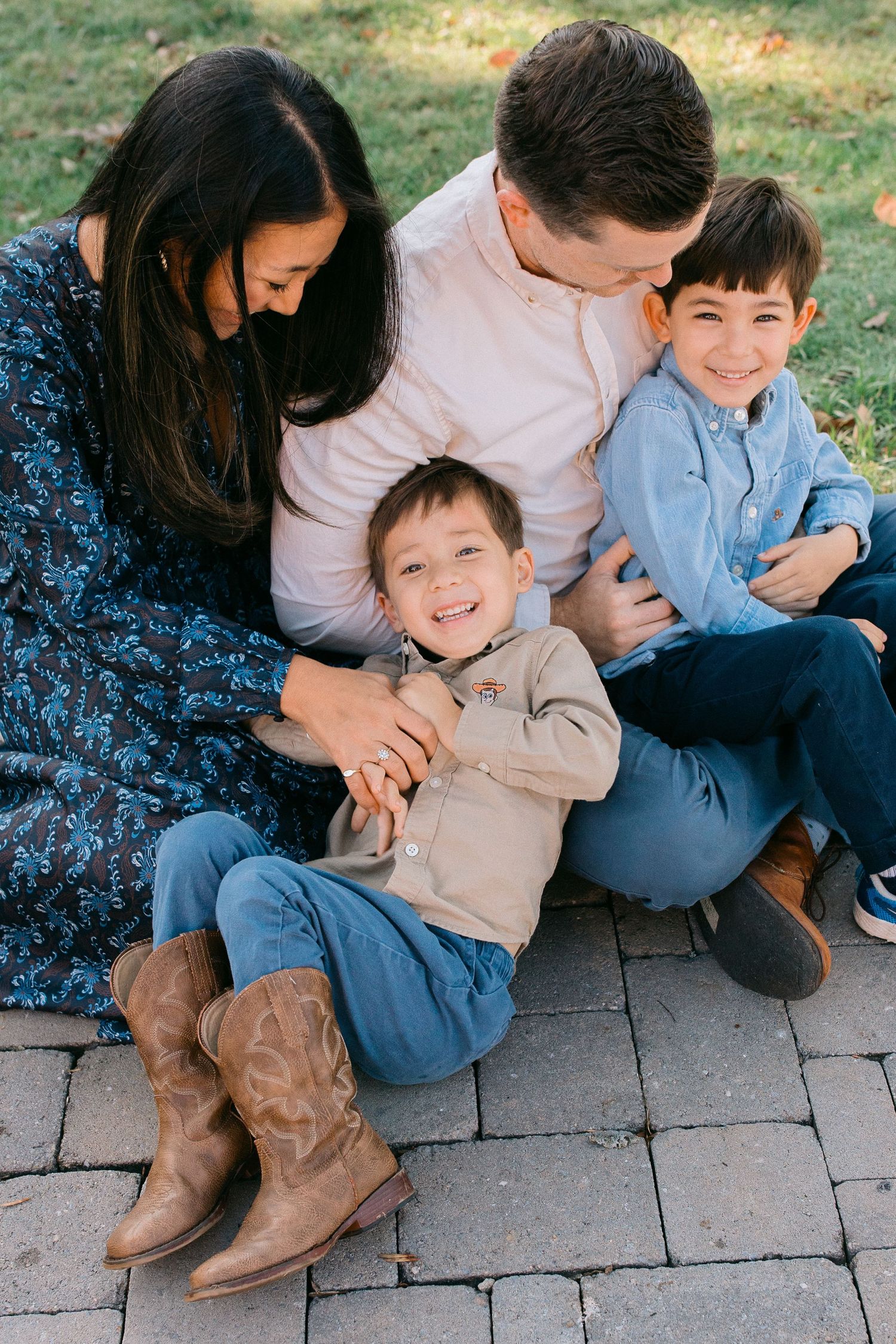 Happy family of four sitting outdoors on brick pavers, parents smiling at two young boys in casual fall attire.