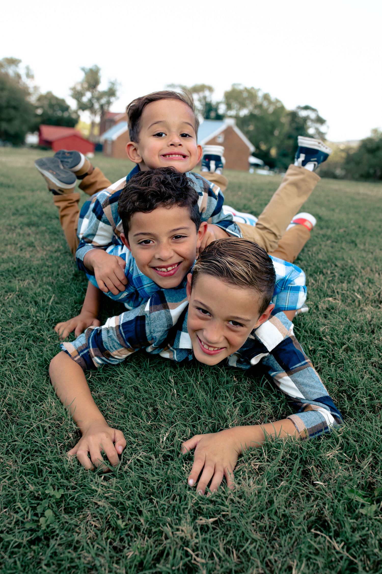 Two young boys in plaid shirts play on green grass while their father watches and smiles in the background.