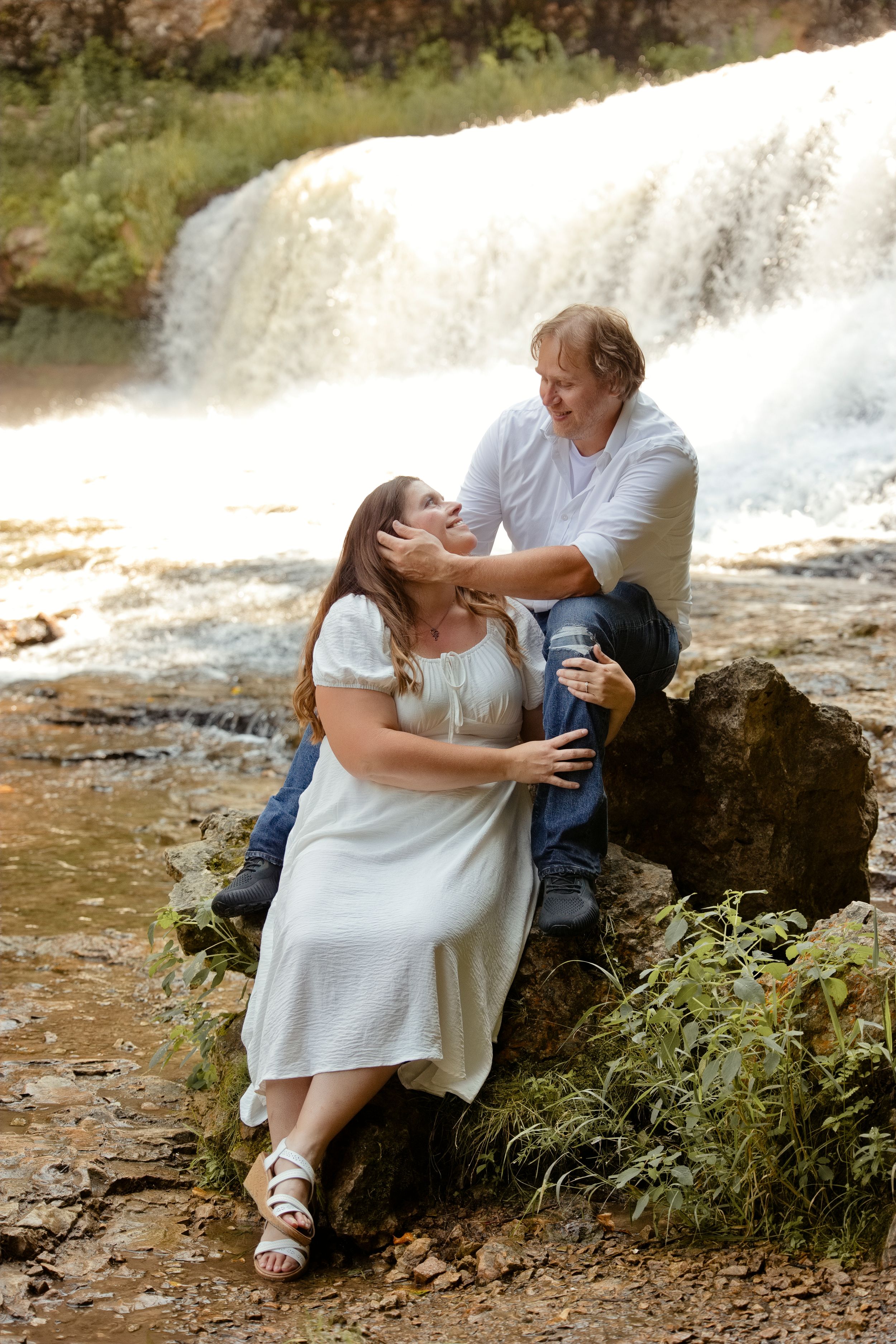 Romantic couple in white outfits sitting on rocks near a waterfall, sharing a tender moment outdoors.