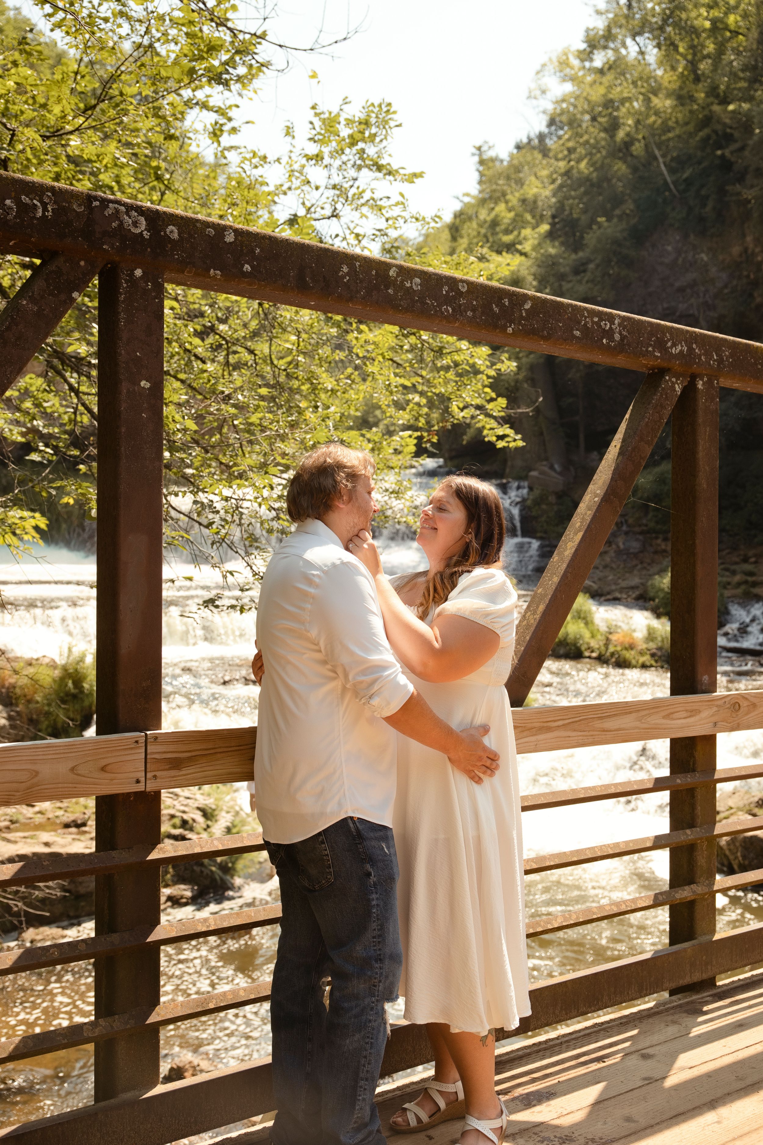 Couple embracing on a wooden bridge with a scenic river and waterfall backdrop on a sunny day.