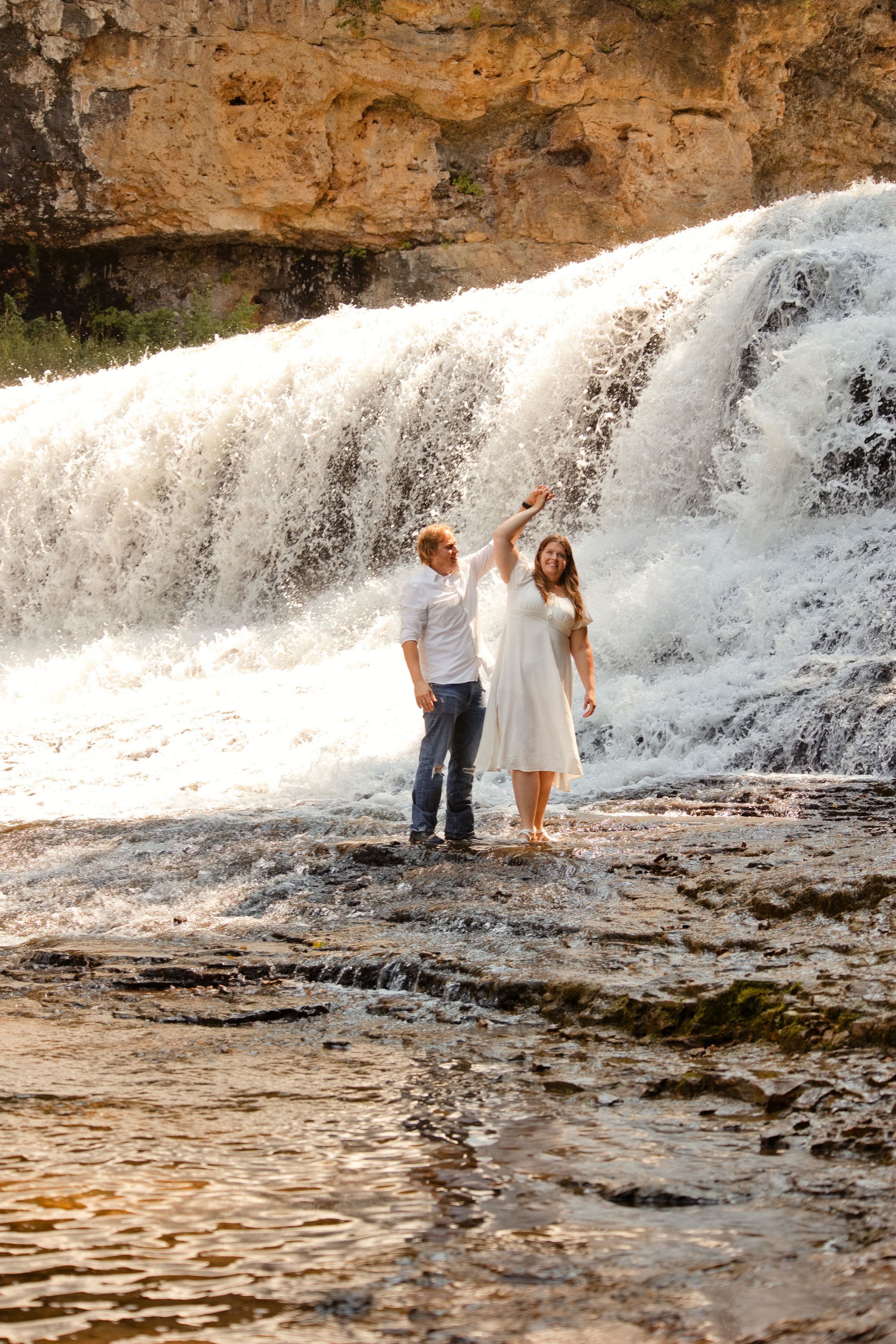Couple holding hands by a stunning waterfall, woman in white dress, man in jeans, standing on wet rocks.