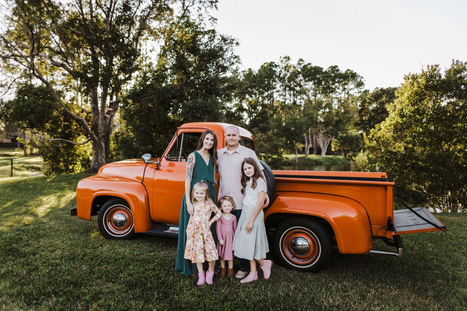 Family of four posing in front of a classic orange vintage pickup truck on a green lawn surrounded by trees.