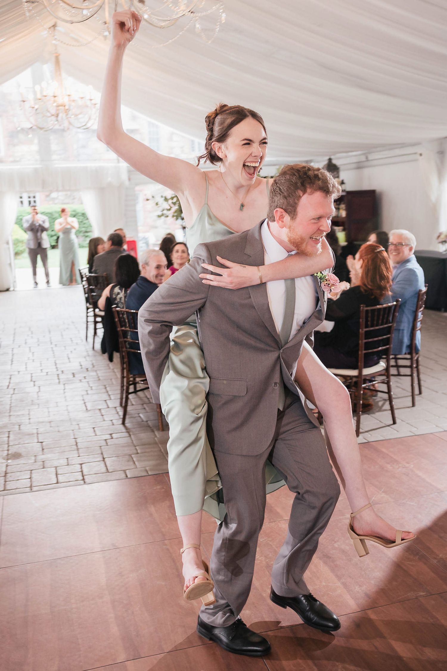 Groomsman carrying a laughing bridesmaid on his back as they enter the reception, guests cheering behind them.