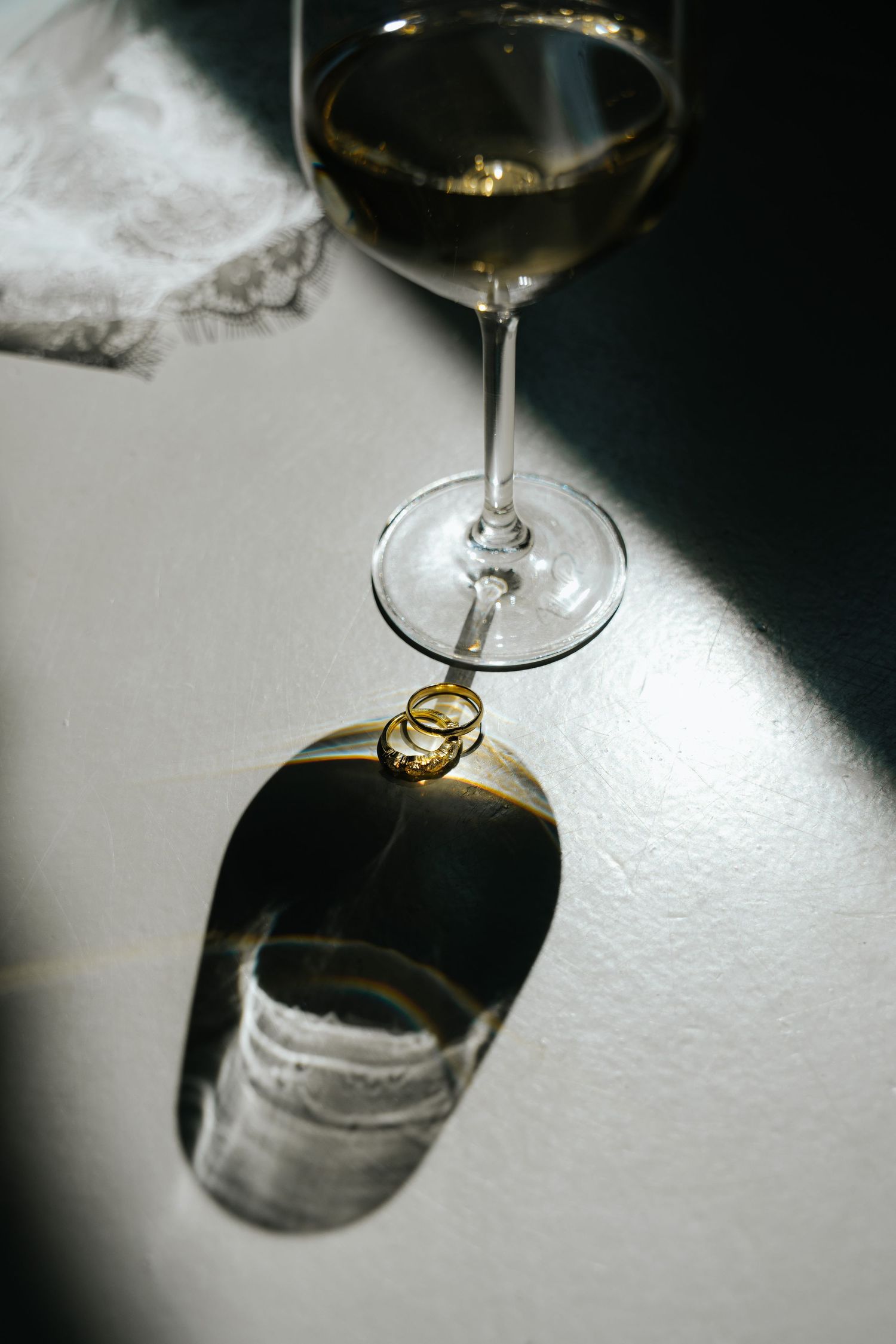 Wedding rings resting against a crystal wine glass casting dramatic shadows on a white surface.
