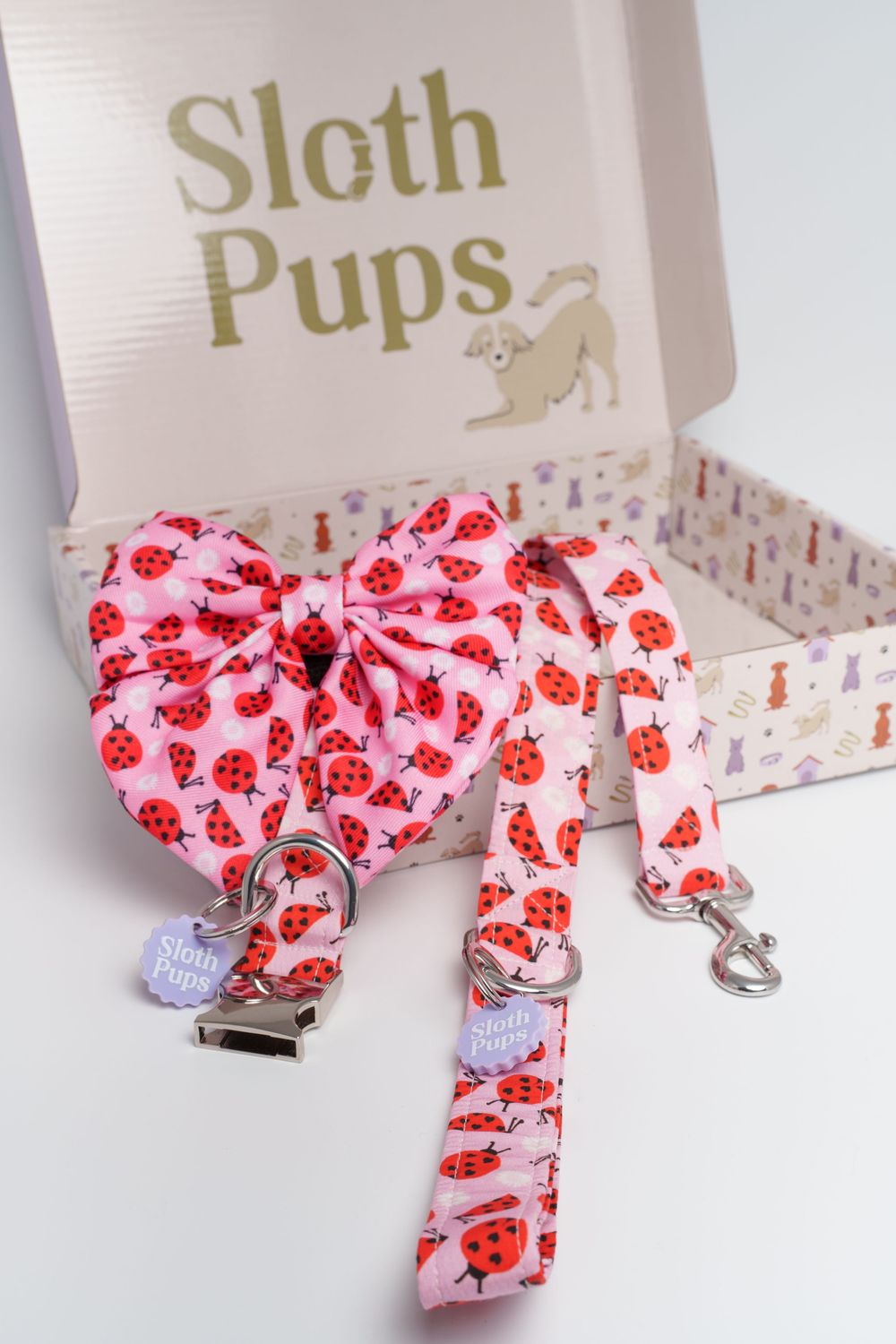 Pink ladybug print dog bow tie, collar, and leash set displayed in a Sloth Pups gift box.