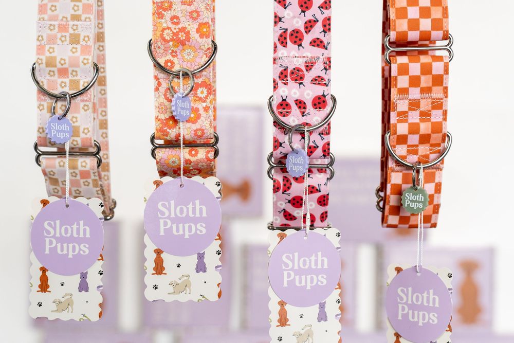 Four colorful Sloth Pups dog collars in floral and checkered patterns displayed with branded tags on a light purple background.