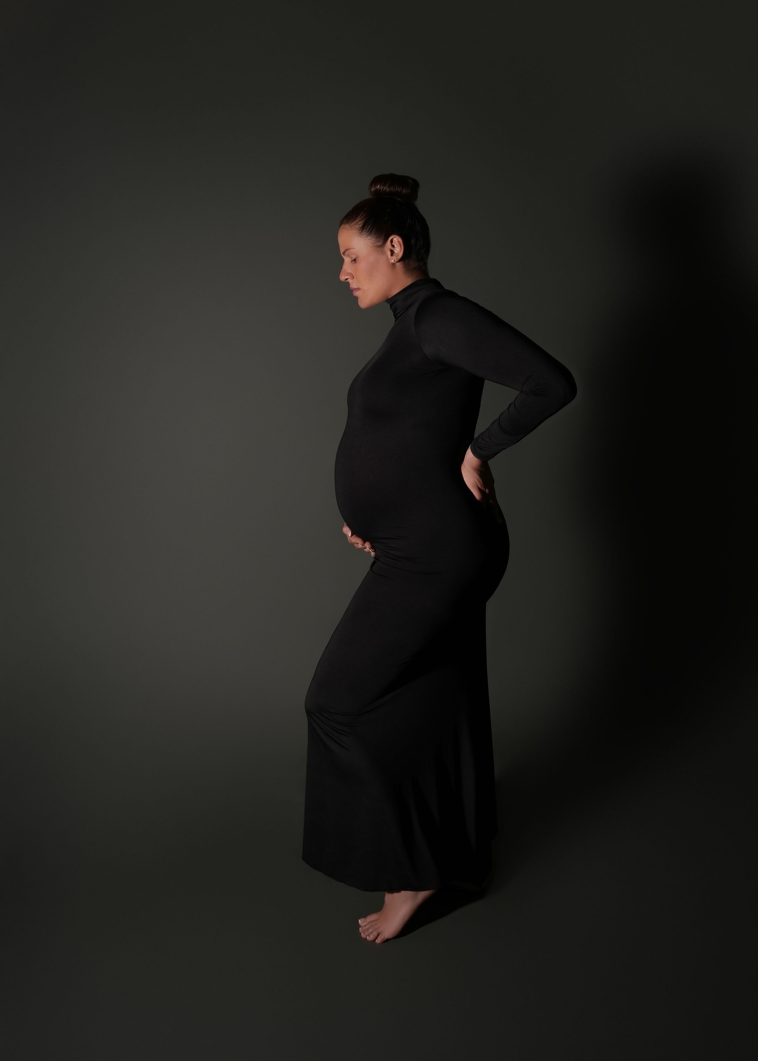 Pregnant woman in black maxi dress poses sideways, cradling baby bump, against dark studio background.