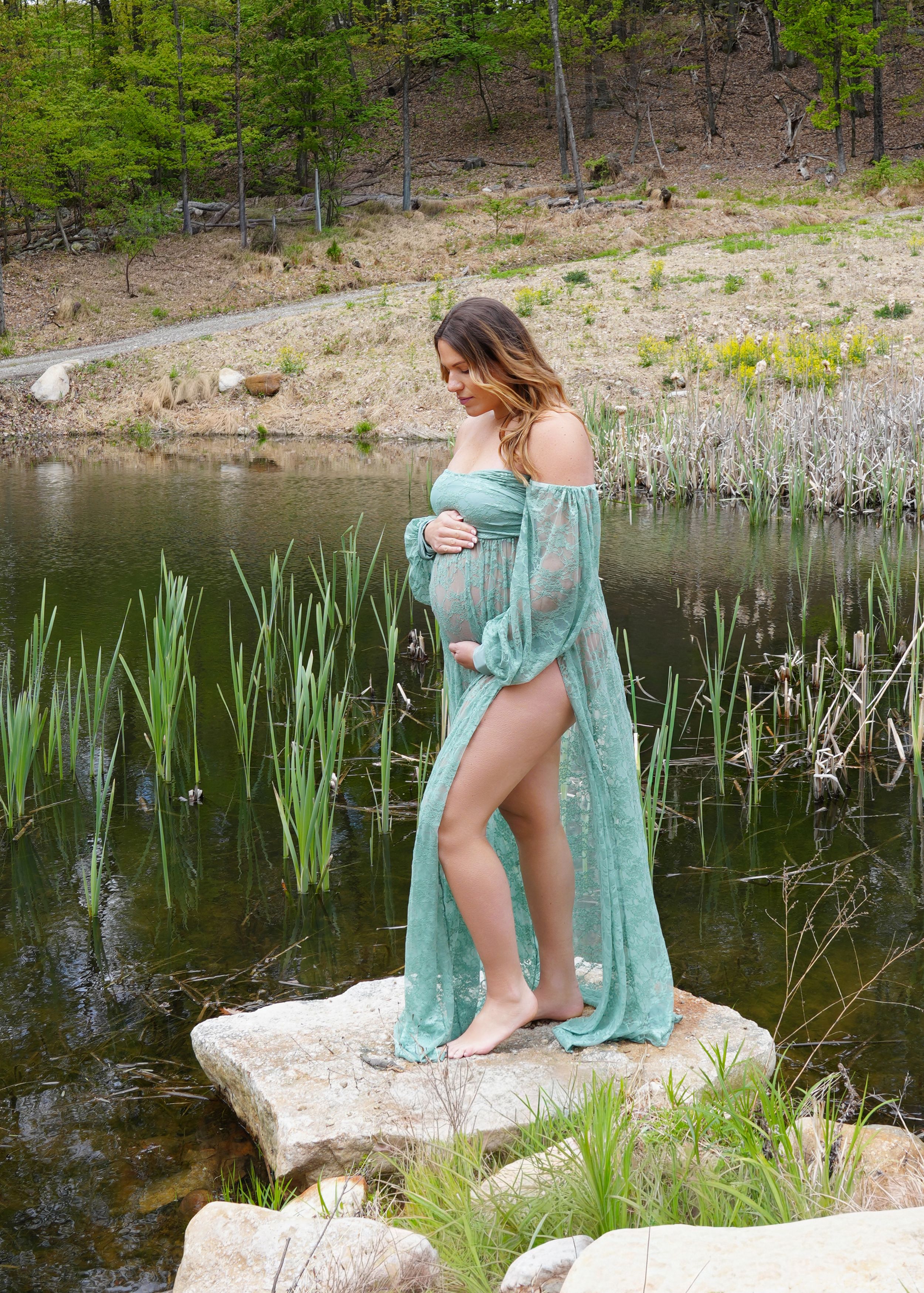 Pregnant woman in teal lace maternity gown standing on rocks by a serene pond surrounded by lush greenery.