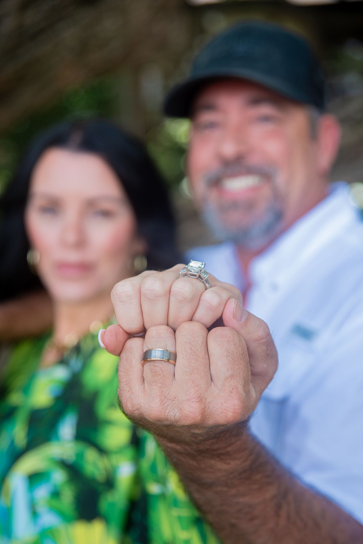 Couple showing off engagement and wedding rings, smiling outdoors, with focus on their hands in the foreground.