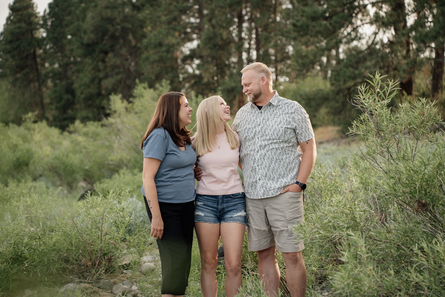 Three people smiling and embracing outdoors near a creek surrounded by lush green trees and shrubs.