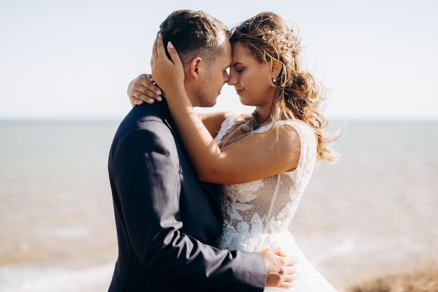 A bride and groom share a romantic moment by the sea, touching foreheads in wedding attire on a sunny day.