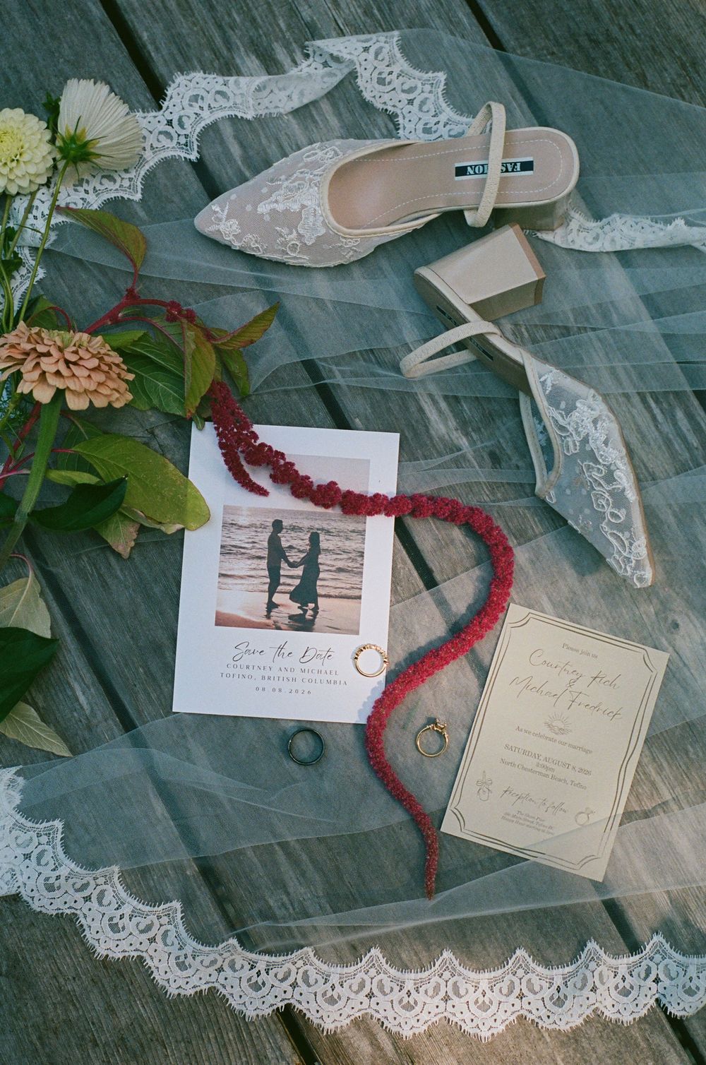 Elegant wedding flat lay featuring lace bridal shoes, rings, red beaded necklace, save the date card, and veil on wood.