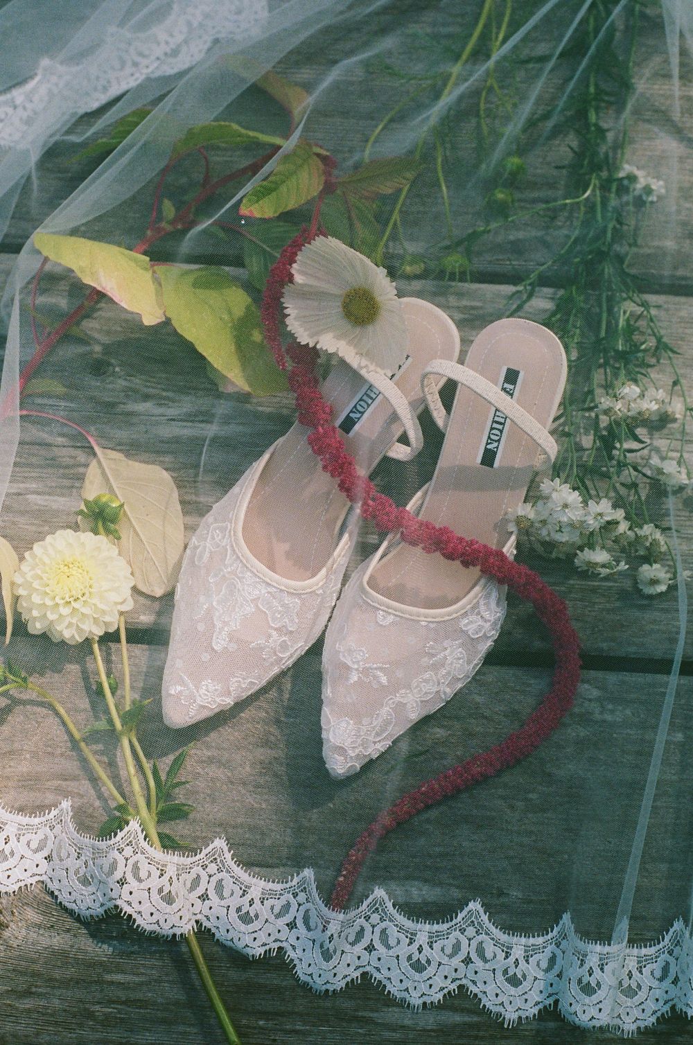 Lace bridal heels surrounded by dahlias, greenery, and a red floral garland on rustic wood.