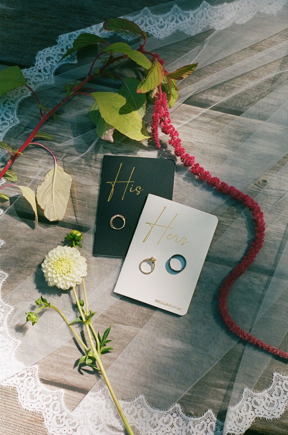 Wedding rings on His and Hers cards with lace veil, white dahlia, and red berry branch on wooden surface.