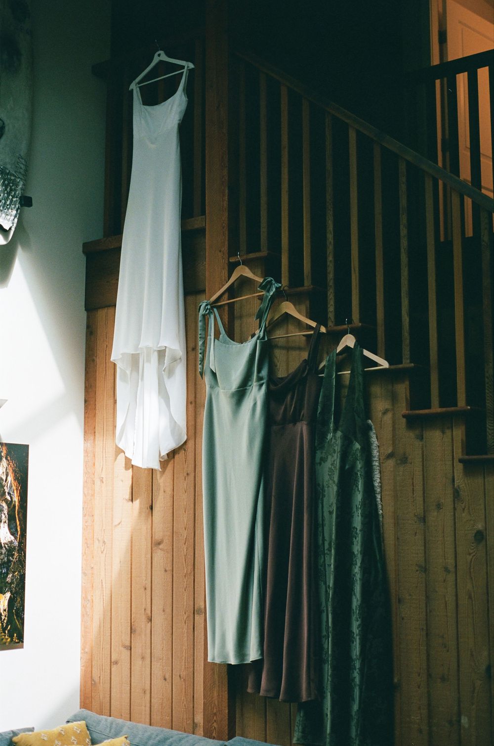 White wedding dress and green bridesmaid dresses hanging on wooden staircase wall in rustic interior.