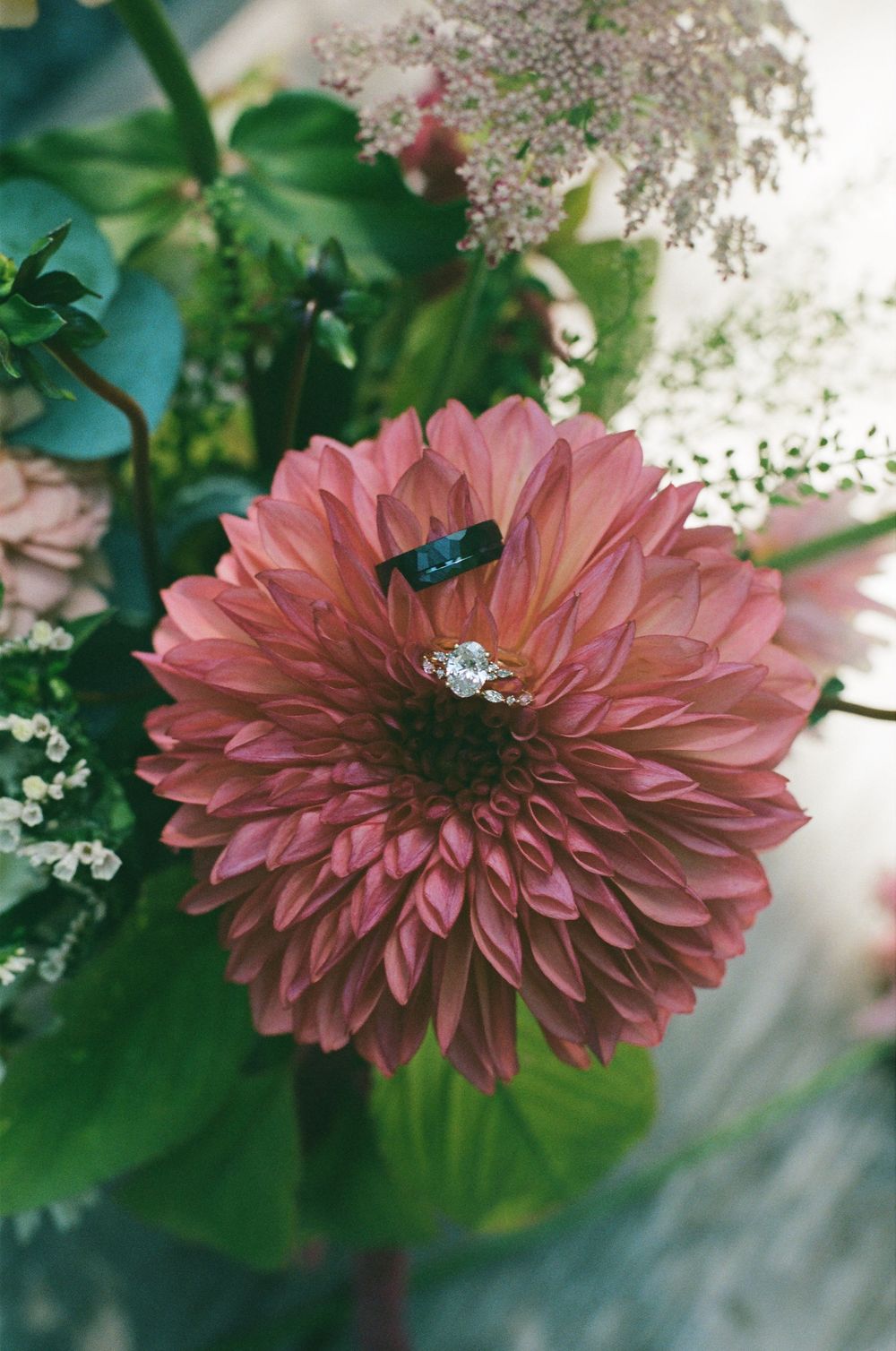 Pink dahlia flower with diamond engagement ring and dark wedding band resting on petals surrounded by lush green floral arrangement.