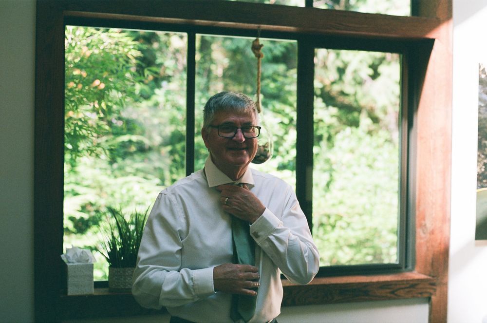 Man in white shirt adjusting green tie near large window with lush green trees in background.