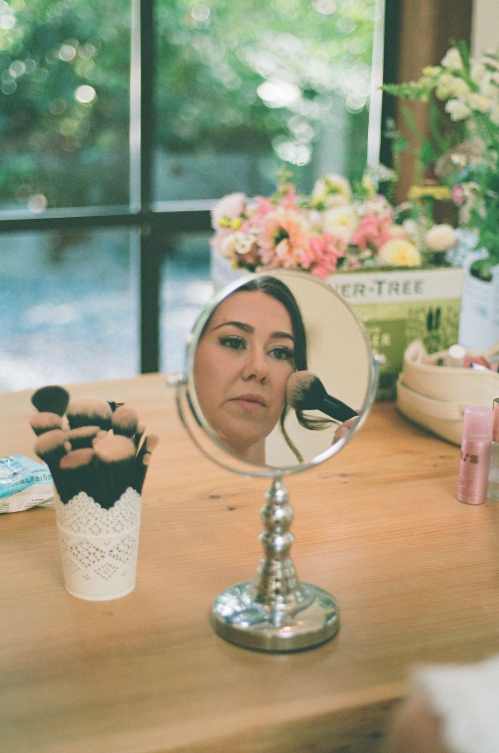 Bride applying makeup while looking into a round vanity mirror on a wooden dressing table.