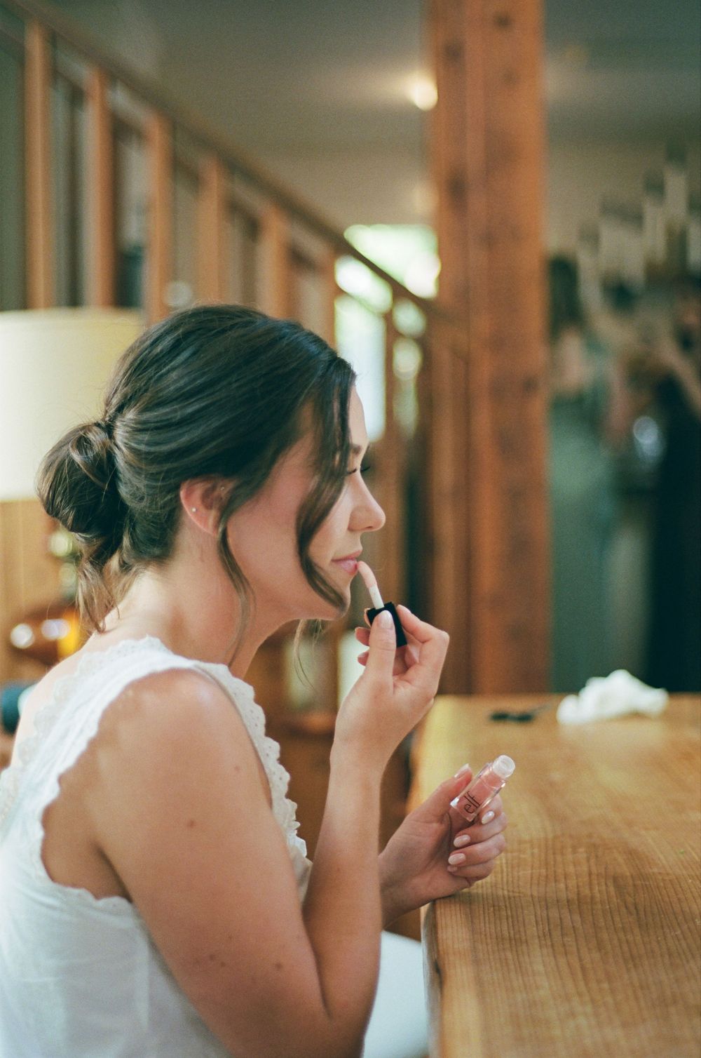 Bride applying lipstick at wooden vanity while getting ready in white dress with delicate neckline detail.