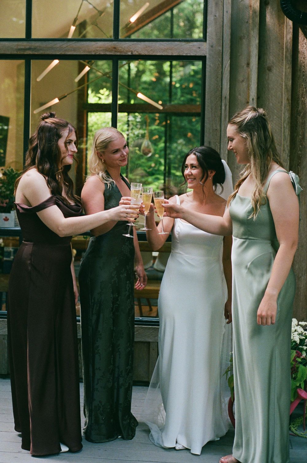 Bride in white gown toasting champagne with three bridesmaids in mismatched dark dresses at rustic venue.