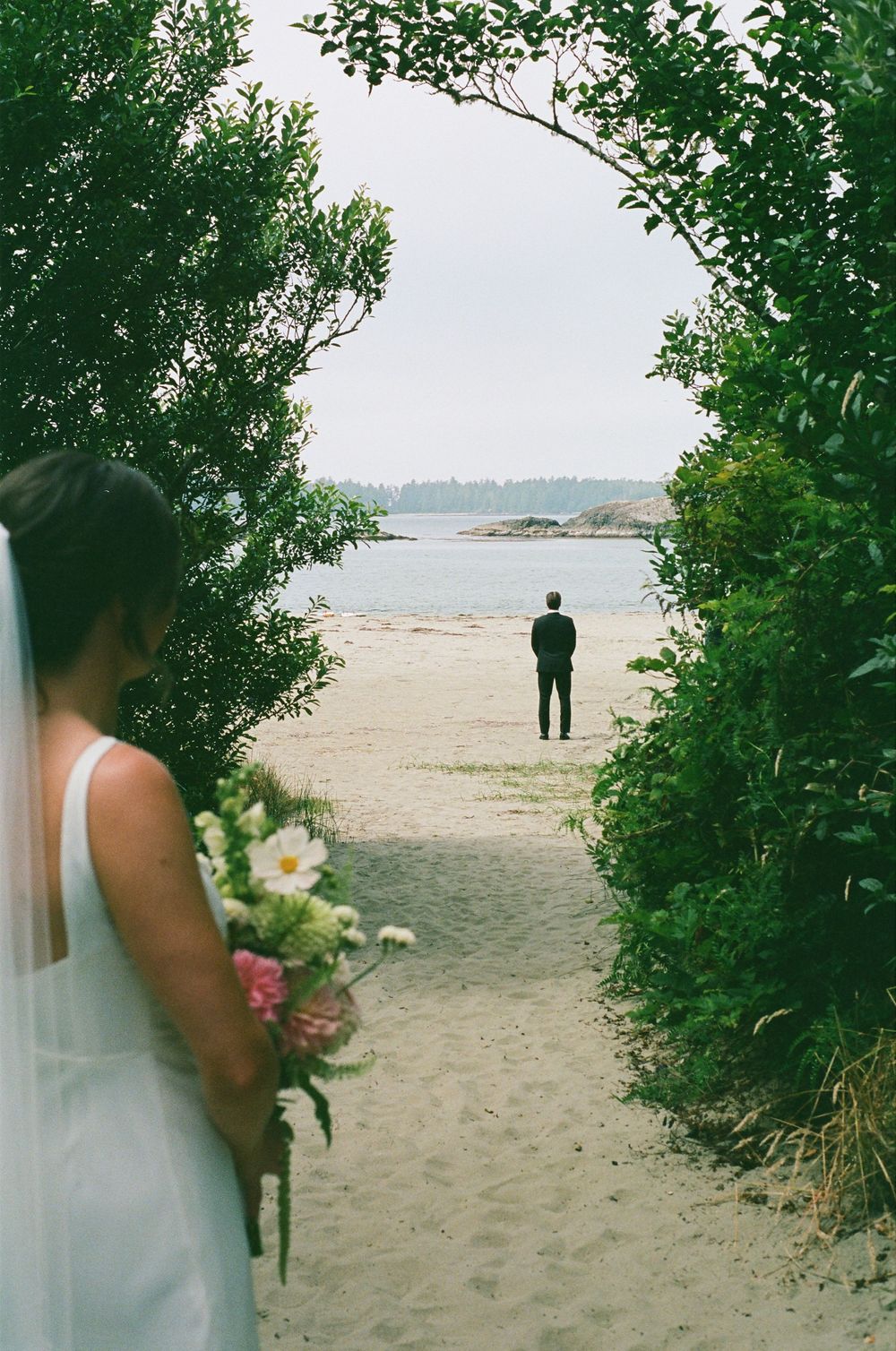 Bride holding bouquet approaching groom standing alone on sandy beach through green archway.