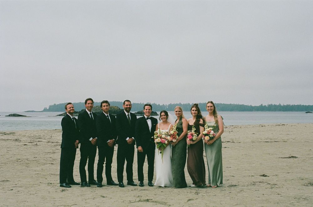 Wedding party poses on a sandy beach, groomsmen in black suits and bridesmaids in green dresses, with ocean backdrop.